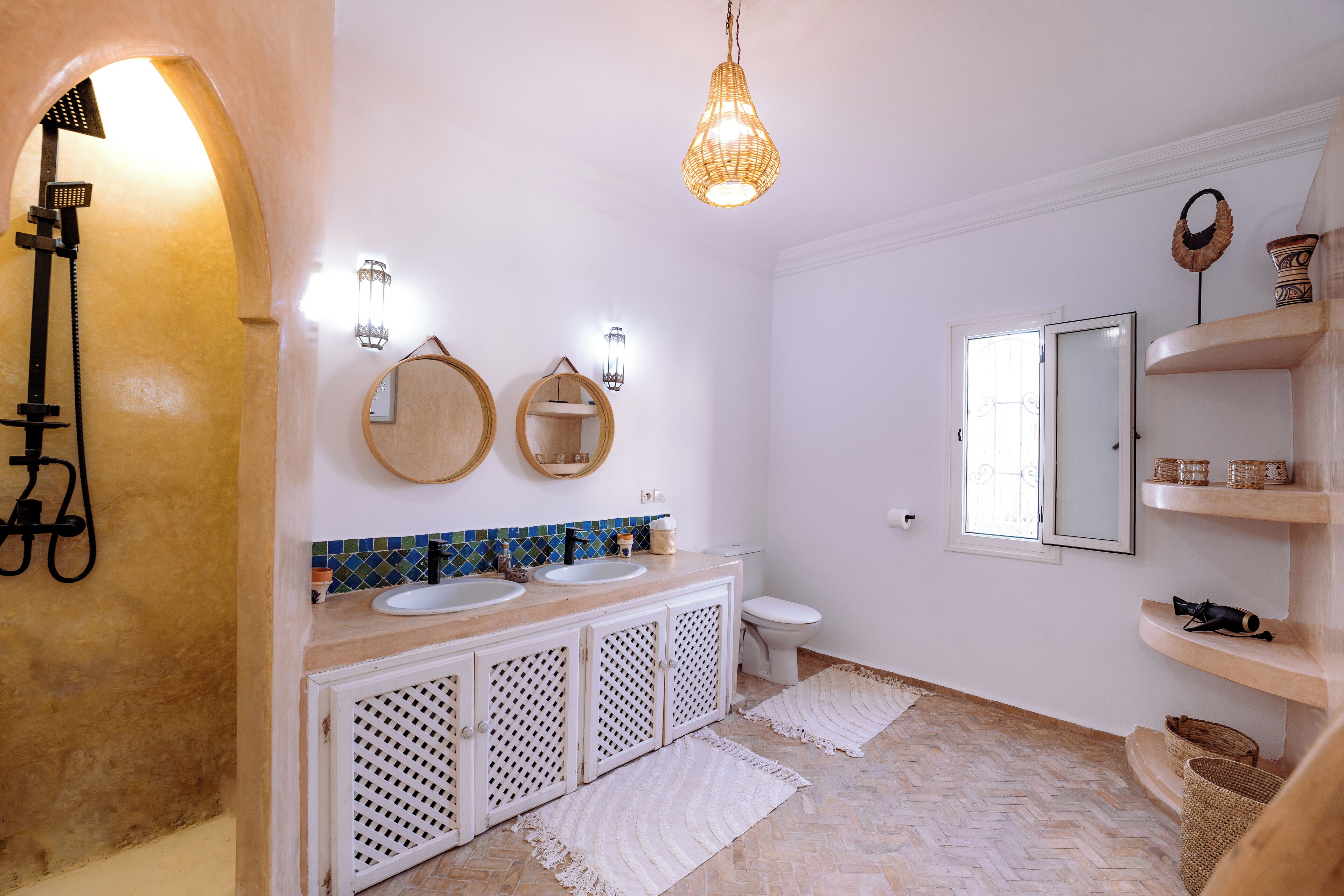 A bathroom with a white vanity, two sinks, a toilet, and a shower. There are wall sconces, a hanging light, and shelves with baskets.
