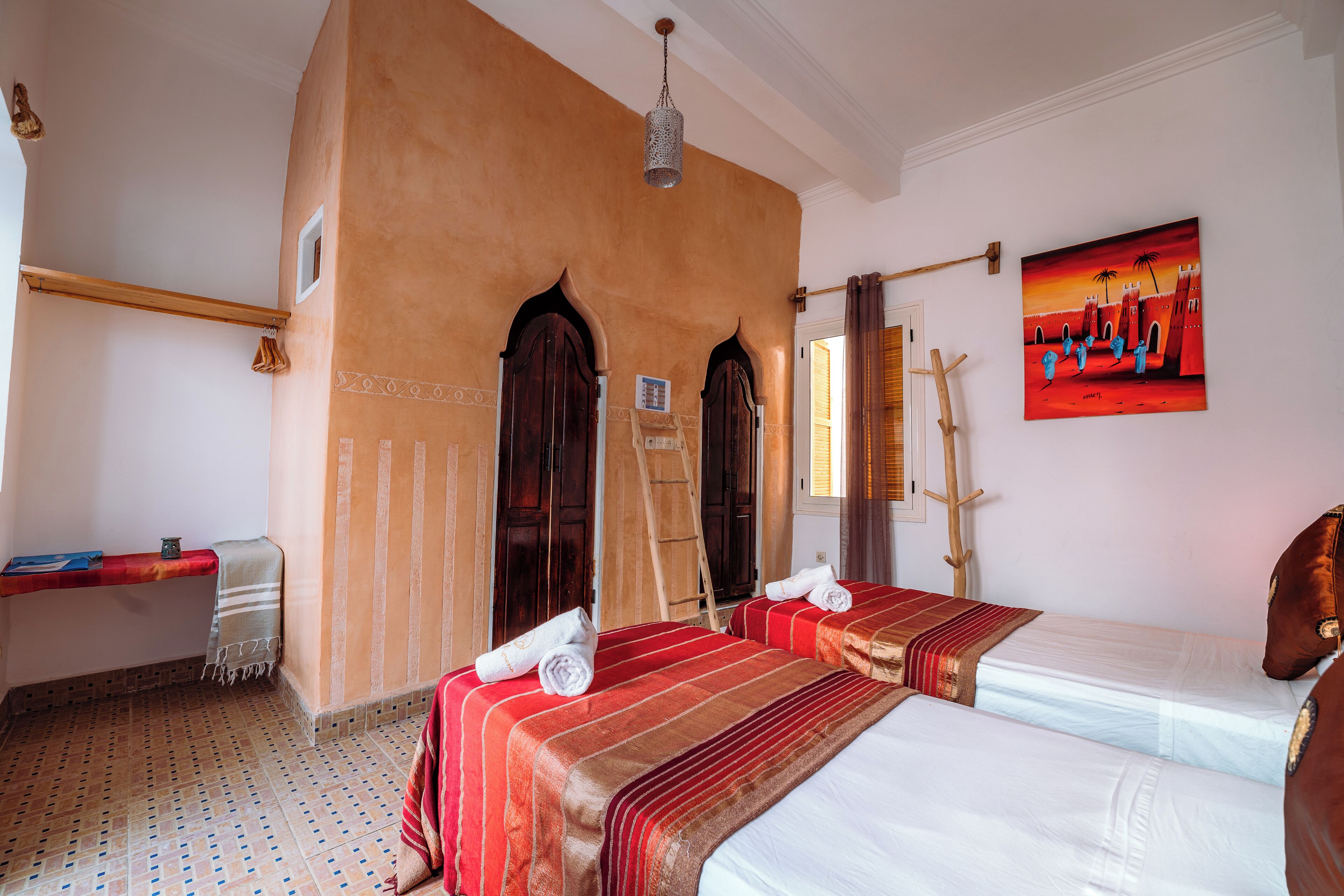 A hotel room with two beds, a wooden ladder, a red and white striped bedspread, and a painting of a sunset scene.
