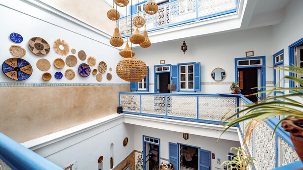 Un interior de varios niveles con ventanas de marco azul, lámparas de mimbre colgantes y platos decorativos en las paredes.