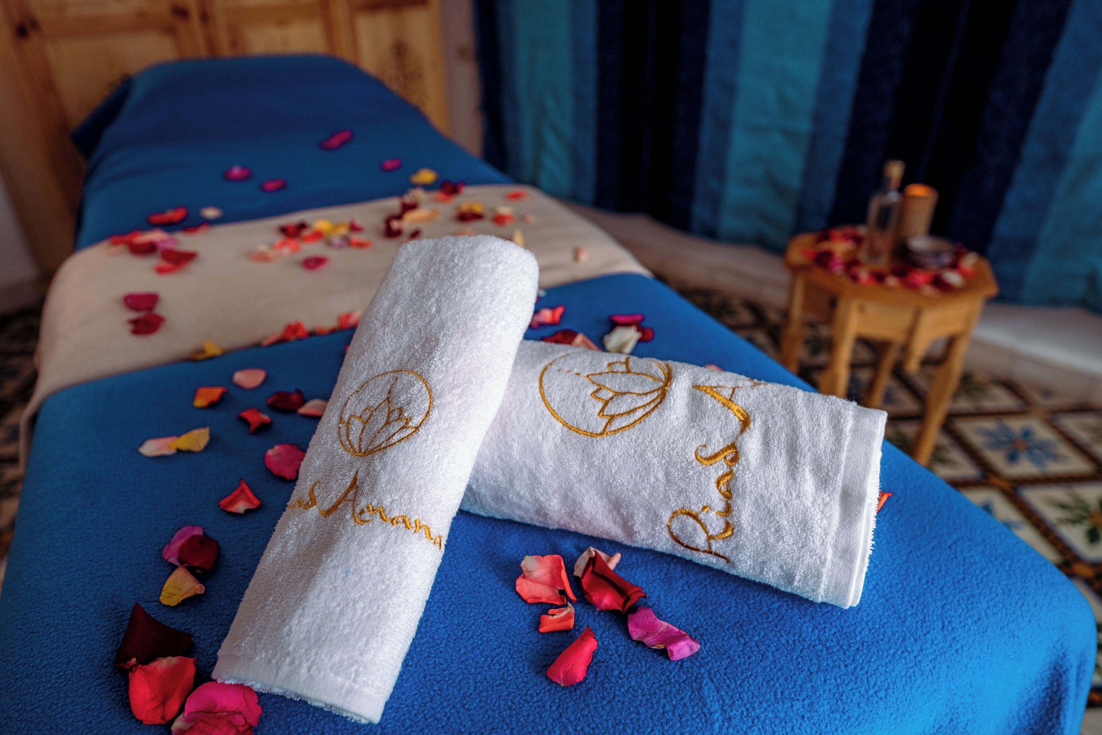 Una cama con sábanas azules y una toalla blanca con texto bordado, rodeada de pétalos de rosa dispersos.