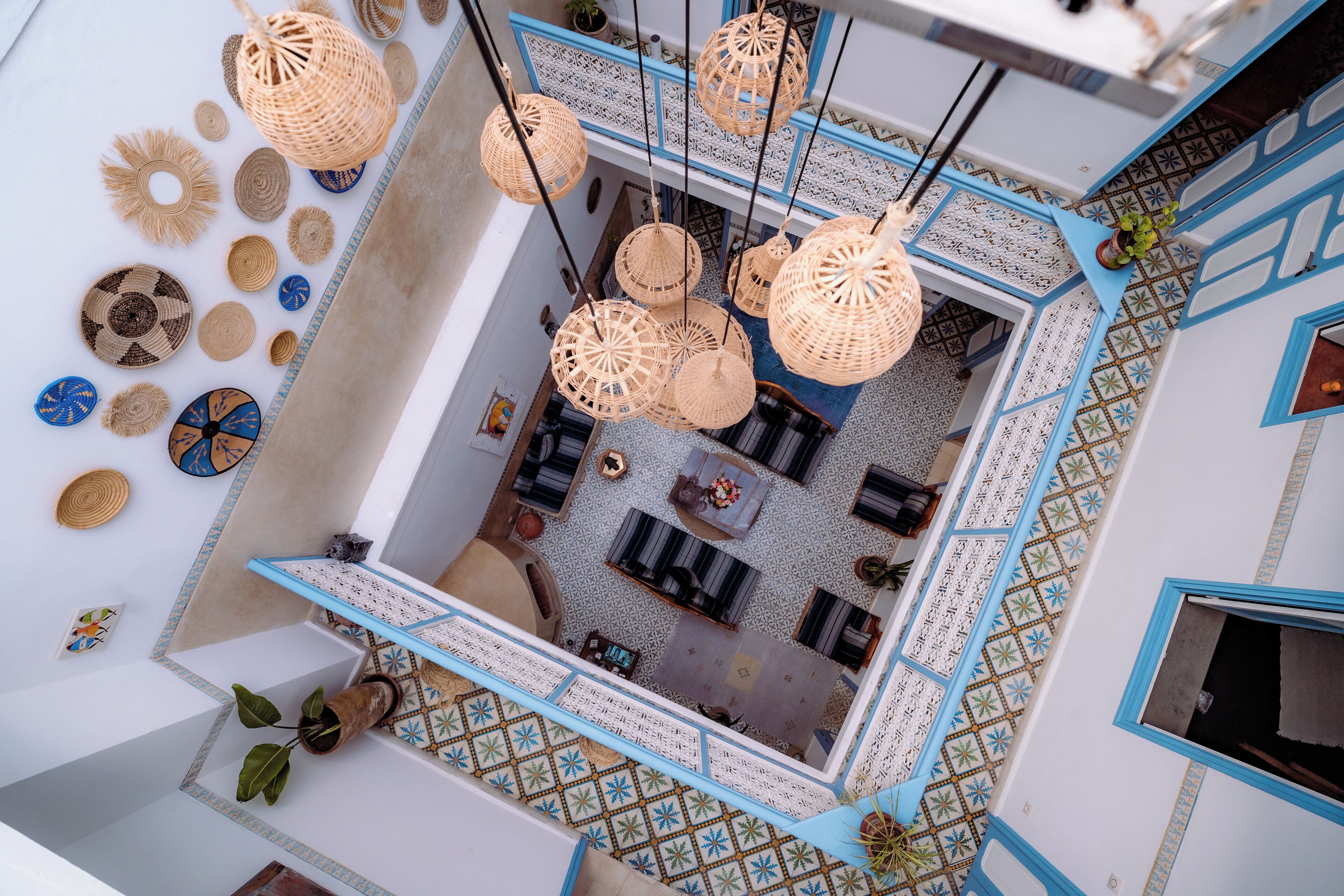 An interior view of a hotel with a central seating area, hanging wicker lamps, and patterned floor tiles.
