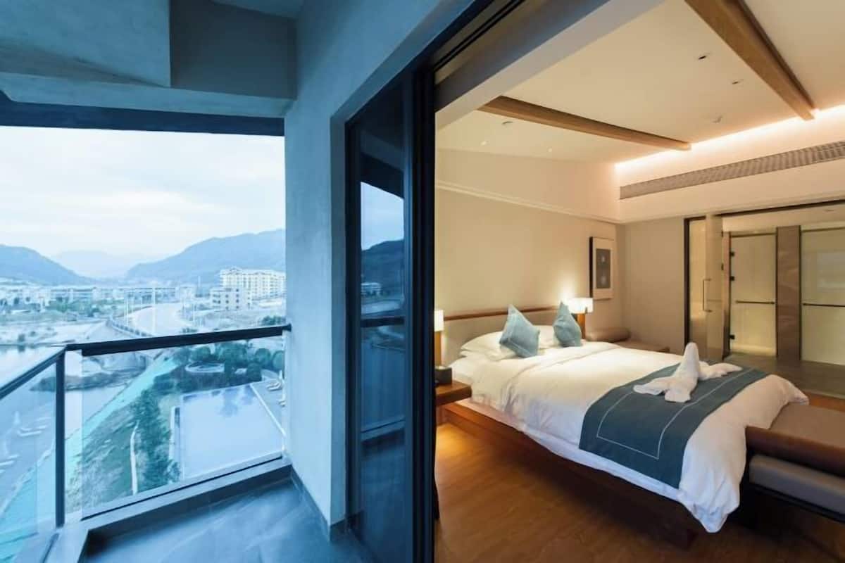 Una habitación de hotel moderna con una cama grande, una mesita de noche y vistas a un paisaje urbano y montañas a través de un ventanal amplio.