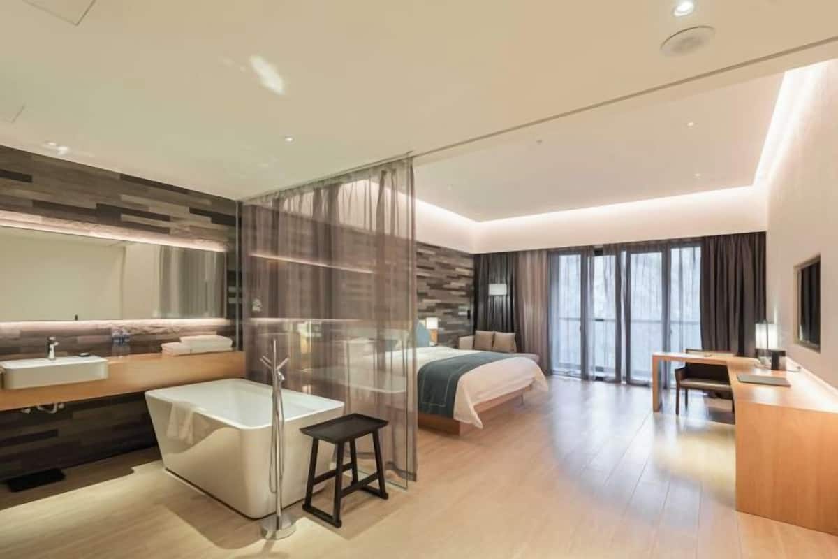 Una habitación de hotel moderna con una cama grande, un baño separado con bañera, un escritorio y un ventanal con cortinas.