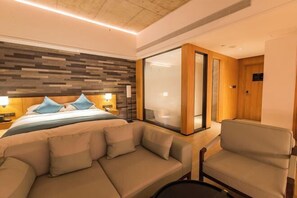 Een hotelkamer met een bed, een slaapbank, een stoel, een houten hoofdeinde en een groot raam.