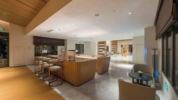 A spacious hotel lobby with a central bar area, seating arrangements, and a view of the kitchen in the background.