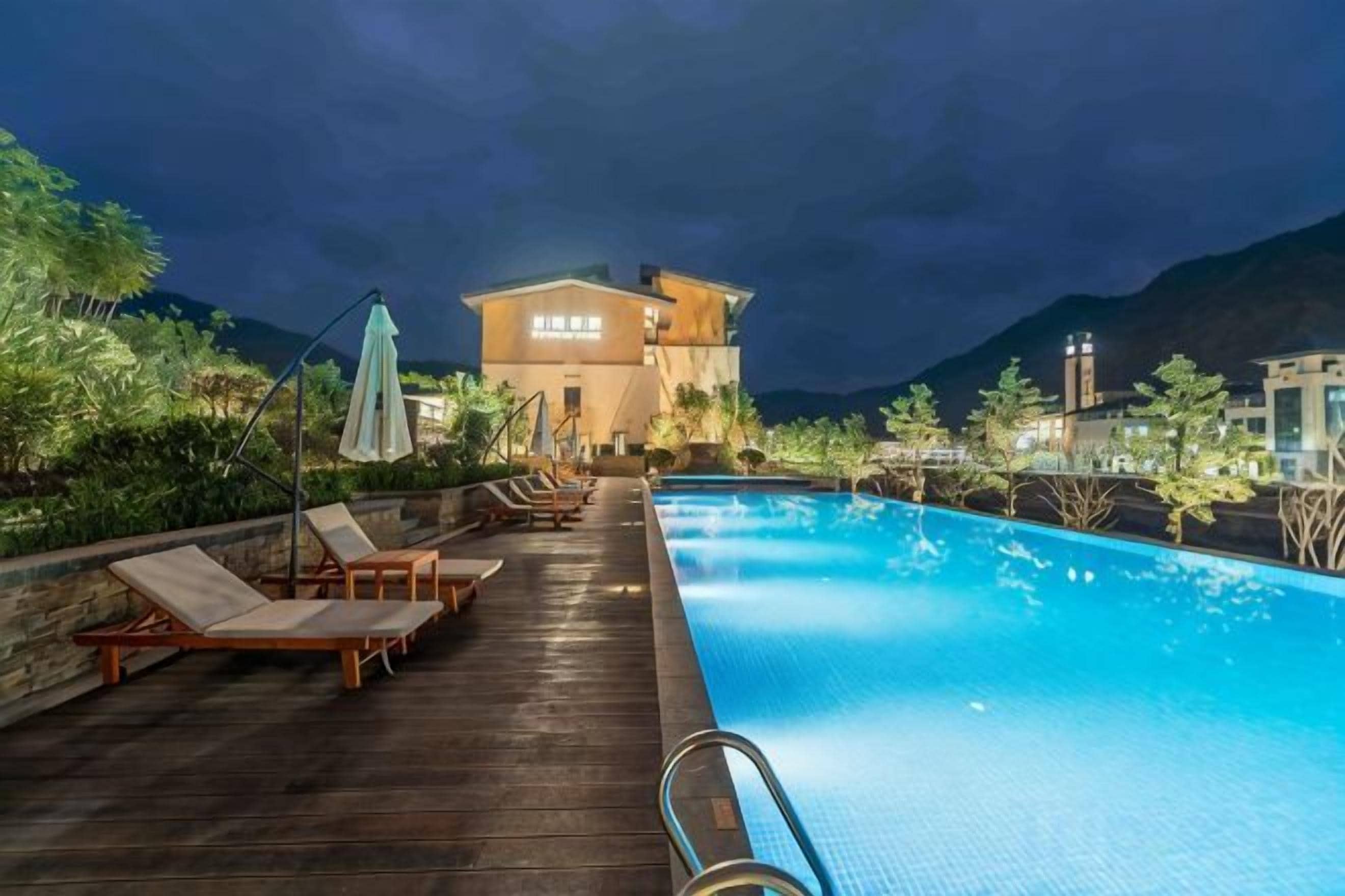 Una piscina con sillones de descanso y sombrillas, una terraza de madera y un edificio con señalización iluminada al fondo.