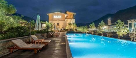 Una piscina con sillones de descanso y sombrillas, una terraza de madera y un edificio con señalización iluminada al fondo.
