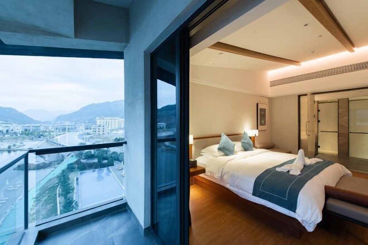 A modern hotel room with a large bed, a bedside table, a nightstand, and a view of a cityscape and mountains through a balcony.