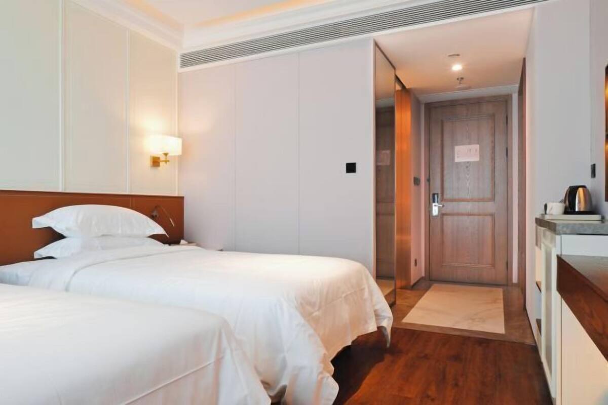 Une chambre d’hôtel avec deux lits, une tête de lit en bois, une lampe fixée au mur et une porte donnant sur une salle de bain.