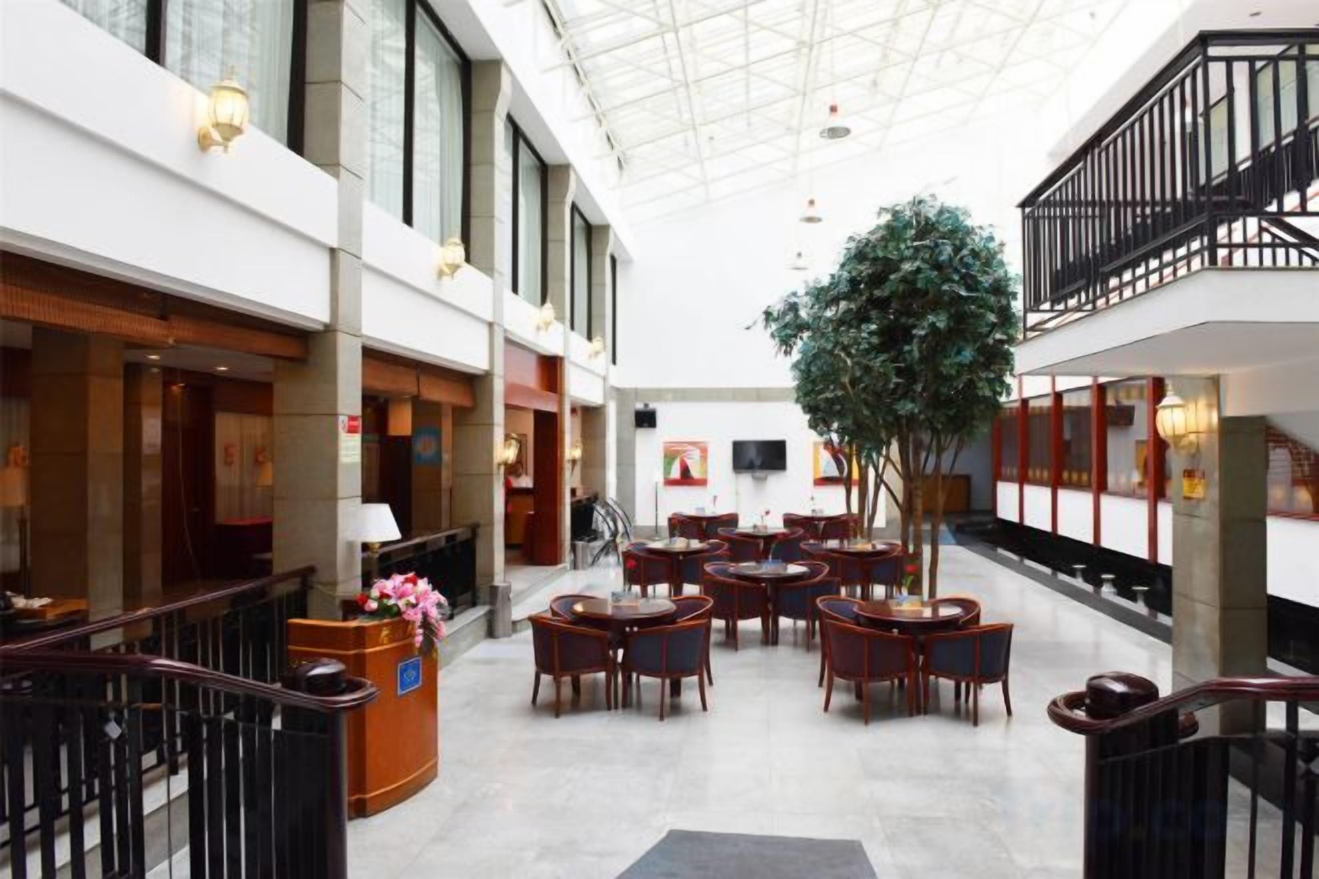 A hotel lobby with a high ceiling, multiple dining tables, a reception desk, and a large indoor plant.