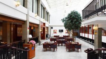 A hotel lobby with a high ceiling, multiple dining tables, a reception desk, and a large indoor plant.