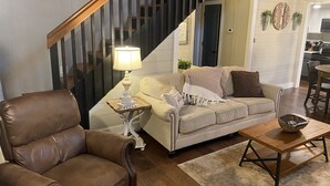 Ett vardagsrum med en brun läderfåtölj, en beige soffa, ett träbord och en lampa på ett sidobord. En trappa med träräcken syns i bakgrunden.