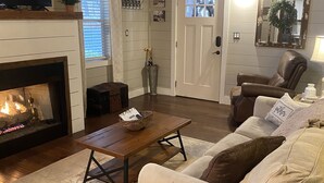 Ett mysigt vardagsrum med en öppen spis, en fåtölj i läder, en soffa och ett soffbord i trä.