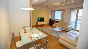 A modern living room with a dining area, a sofa, a TV, and a window with blinds. - Unique Apartment Platnik (Skopje)