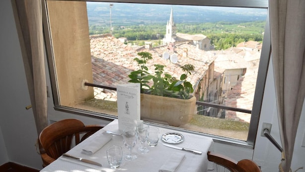 A dining table set with a menu, glasses, and cutlery, overlooking a cityscape with a church spire. - Caesar Panoramic Hotel Restaurant (Bonnieux)