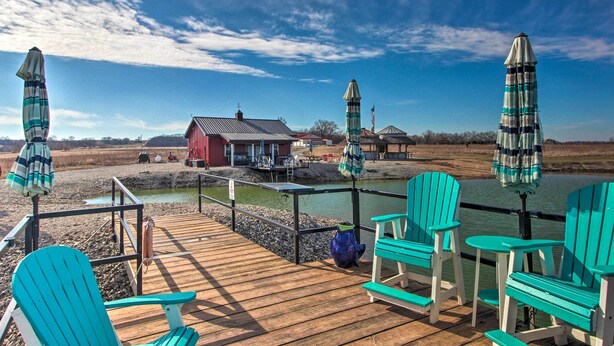A wooden deck with turquoise Adirondack chairs and striped umbrellas, overlooking a river and a red building. - Pheasant Game Farm Missouri Vacation Rental! (Braymer)