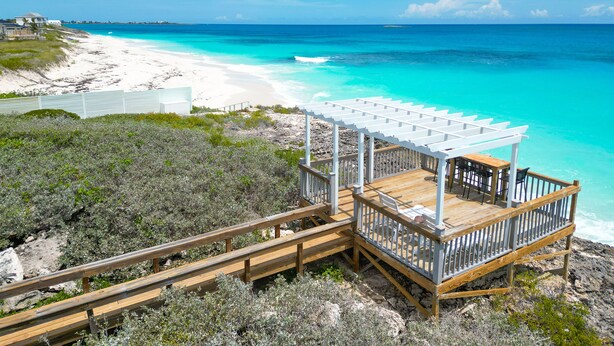 Eine Holzterrasse mit einer Pergola mit Blick auf einen Strand und klares türkisfarbenes Wasser.