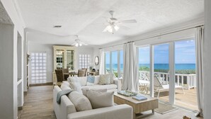 Ein geräumiges Wohnzimmer mit großem Fenster zum Meer, Deckenventilator und einer gemütlichen Sitzecke.