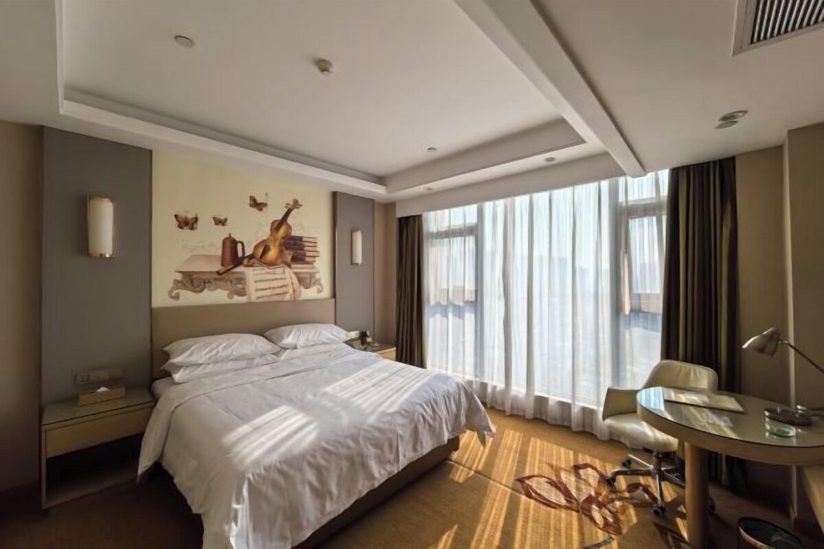 A hotel room with a bed, a desk, a chair, a lamp, and a large window with curtains.