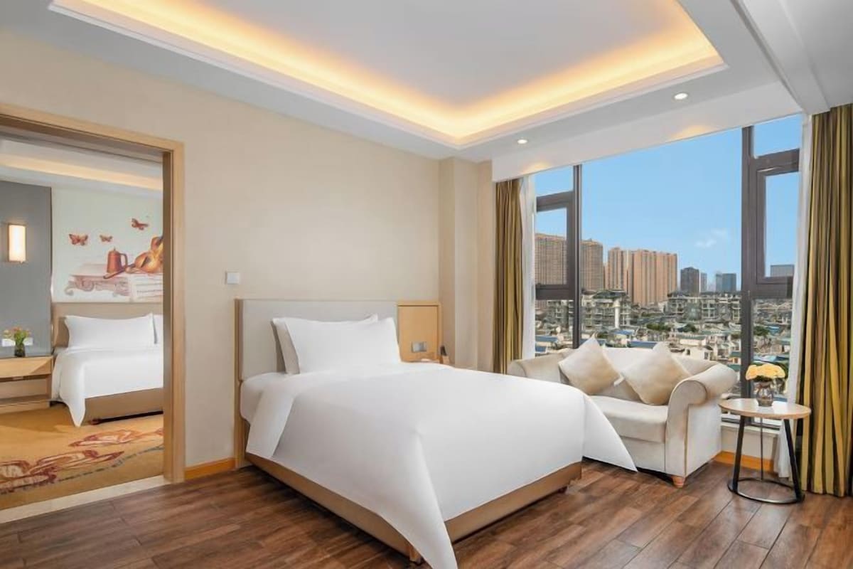 A modern hotel room with a large bed, a sofa, and a view of a cityscape through large windows.
