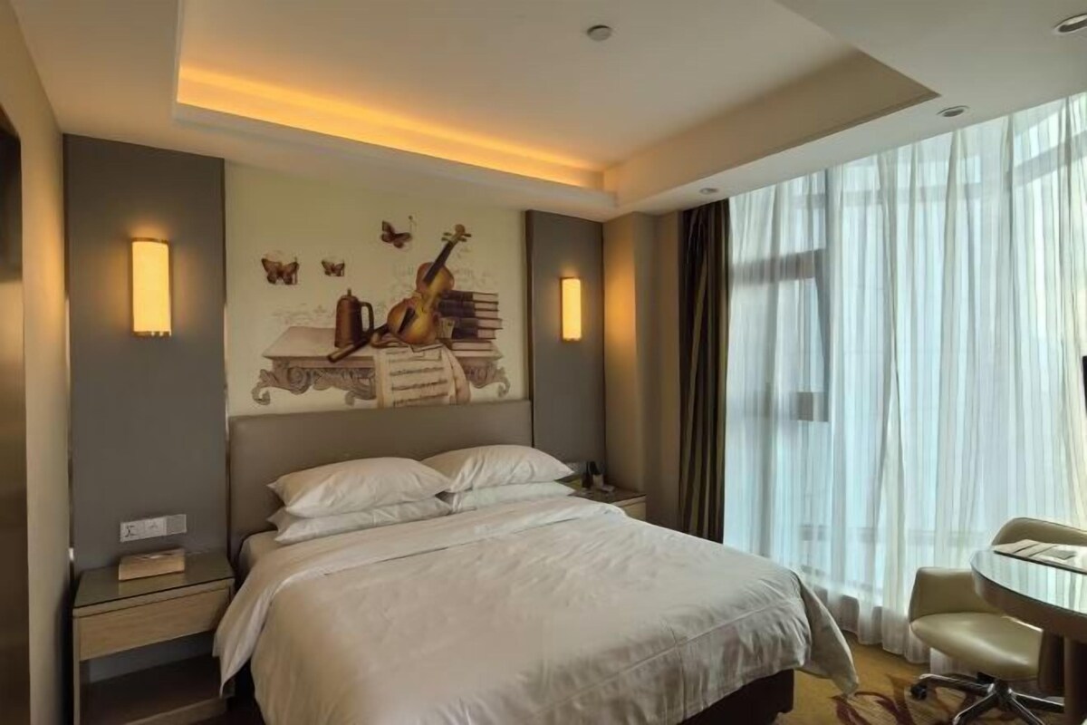 A hotel room with a bed, bedside tables, a desk, a chair, and a large window with curtains.