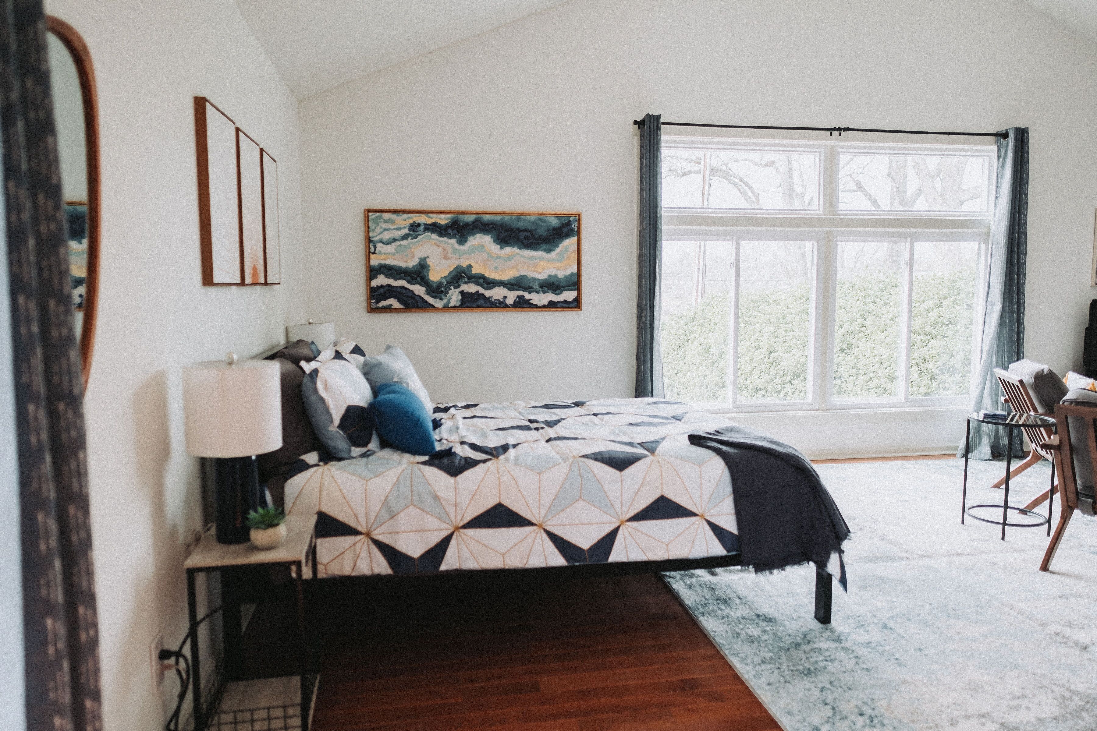 A bedroom with a bed, a desk, a chair, a window, and a painting on the wall.