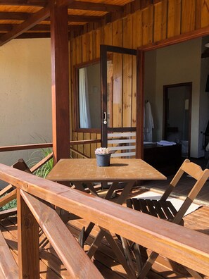 Una terraza de madera con una mesa, sillas, una planta en maceta y una puerta de madera que da a un espacio interior.