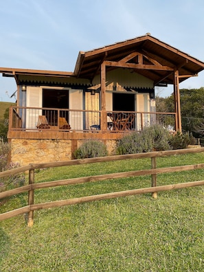 Una cabaña rústica con terraza de madera y cimientos de piedra, rodeada de una cerca de madera y follaje.