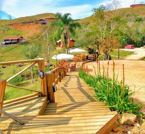 Una terraza de madera que da a un patio con mobiliario exterior y sombrillas, rodeado de vegetación y colinas al fondo.
