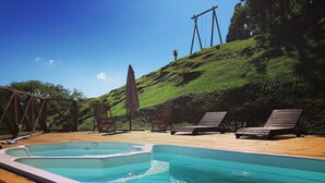 Une piscine entourée d’une terrasse en bois et d’une colline herbeuse, avec des chaises longues et un parasol à proximité.