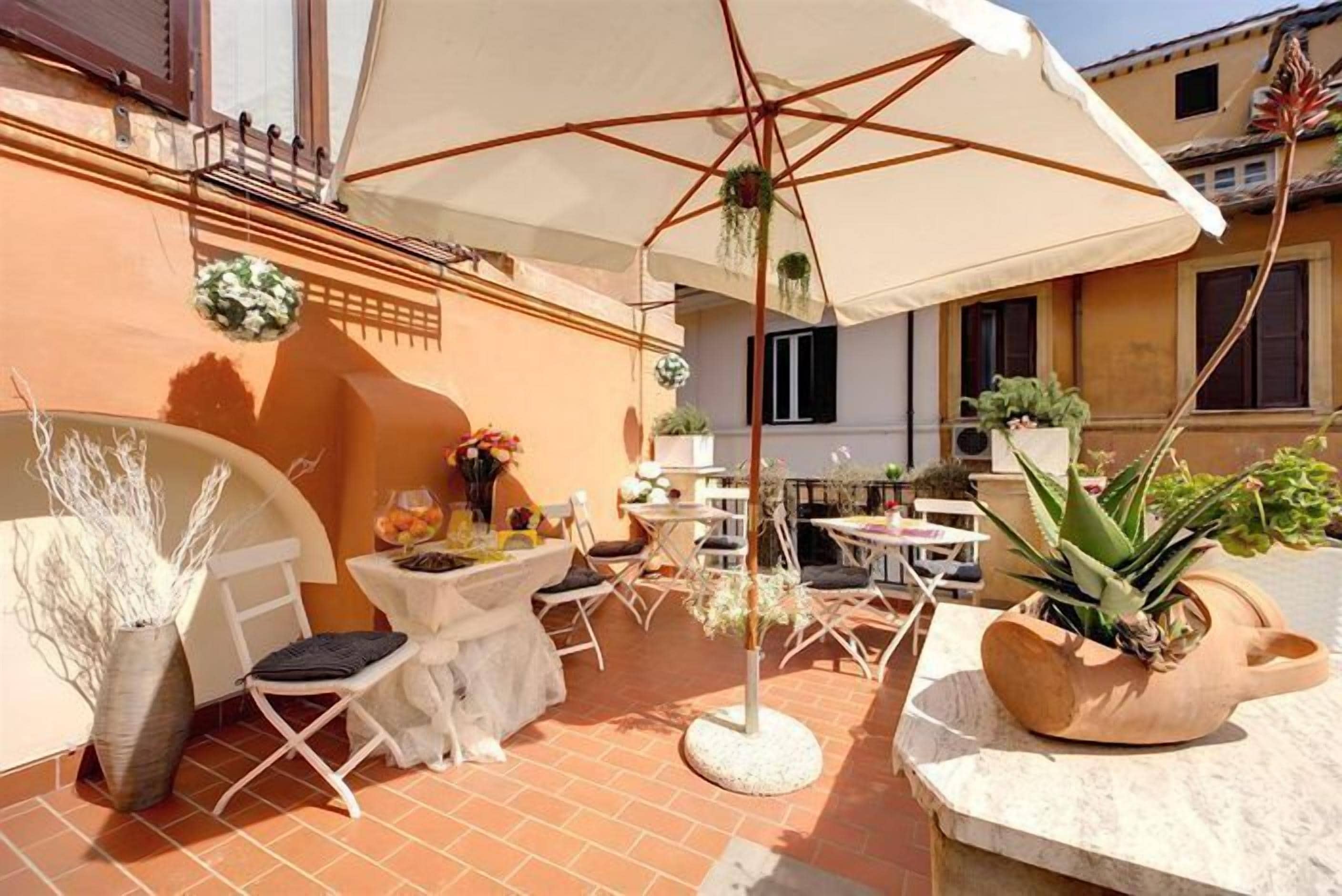 Un patio al aire libre con sillas y mesas blancas, un paraguas grande y plantas en macetas.