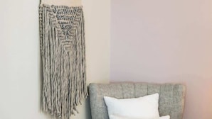 Un fauteuil recouvert d’un tissu gris, agrémenté d’un coussin blanc, une décoration murale en macramé et un plancher en bois.
