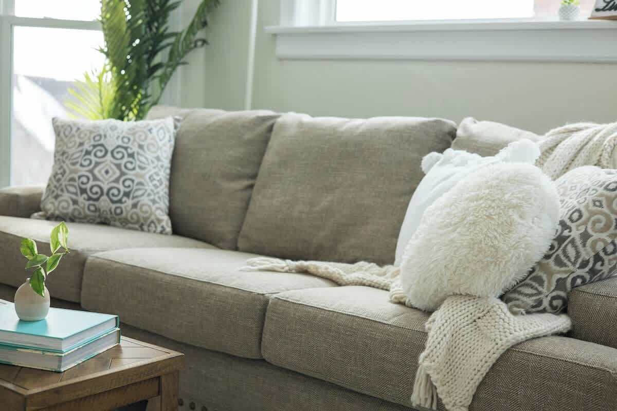 Un canapé beige agrémenté de coussins à motifs, un plaid blanc en tricot et une petite plante posée sur une table d’appoint en bois.