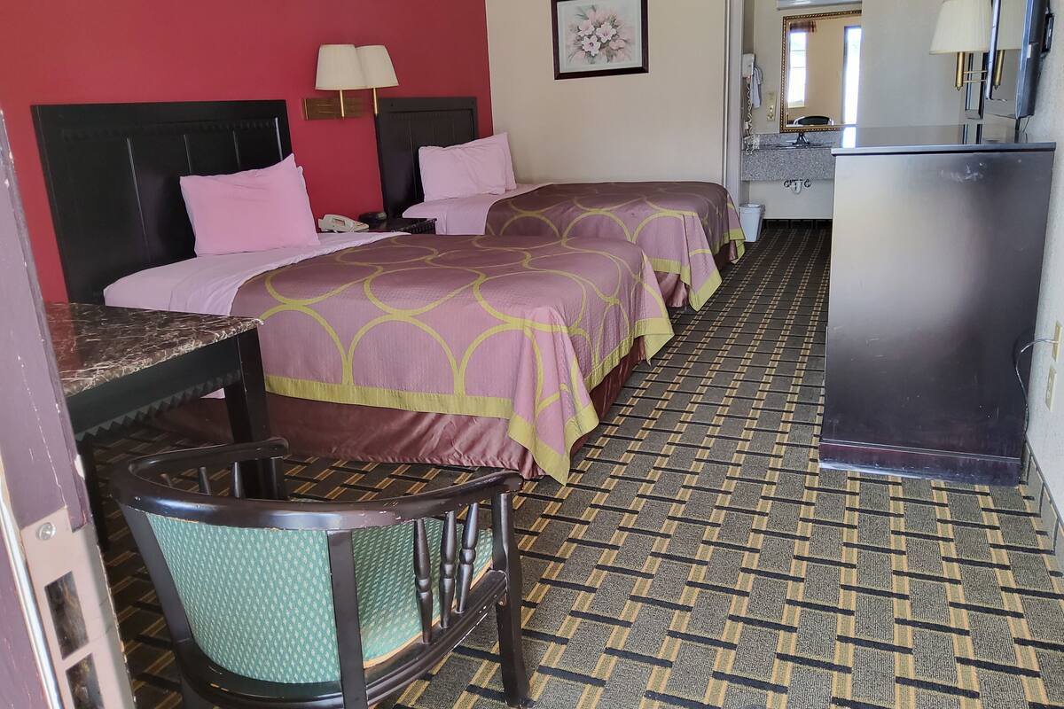A hotel room with two beds, a desk, a chair, a desk lamp, a mirror, and a refrigerator.