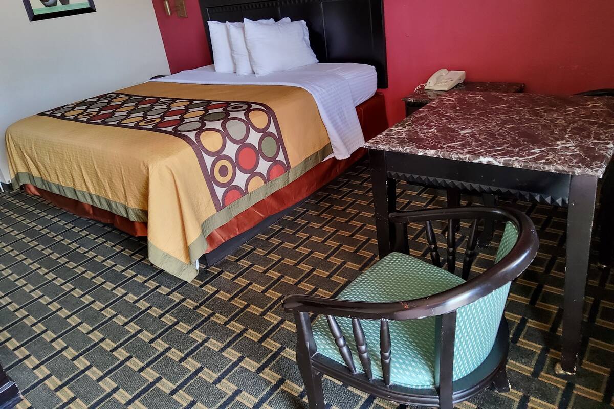 A hotel room with a bed, a table, a chair, a telephone, and a wall with a framed picture.