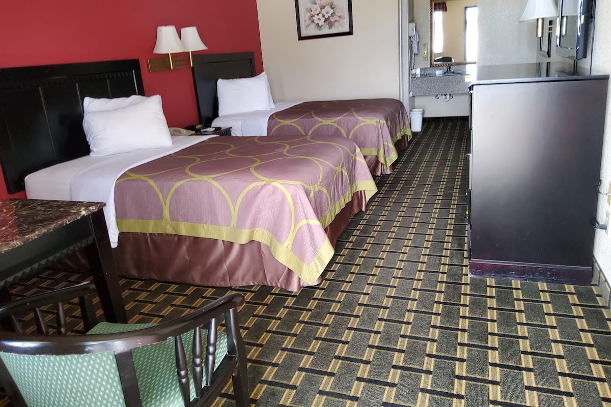 A hotel room with two beds, a desk, a chair, a refrigerator, a bathroom with a sink and mirror, and a patterned carpet.