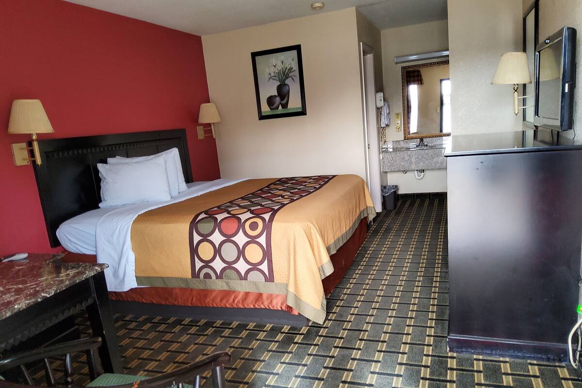 A hotel room with a bed, a desk, a chair, a television, and a bathroom visible in the background.