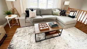 A living room with a sectional sofa, a coffee table with board games, and a view of the outdoors through large windows. - Upper Unit 1067 Rawding  (Port Williams)