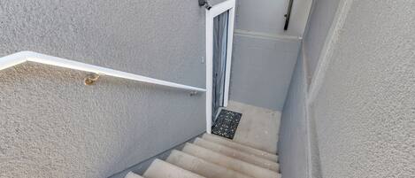 Un escalier étroit avec une rampe blanche, une porte en haut et un petit luminaire décoratif.