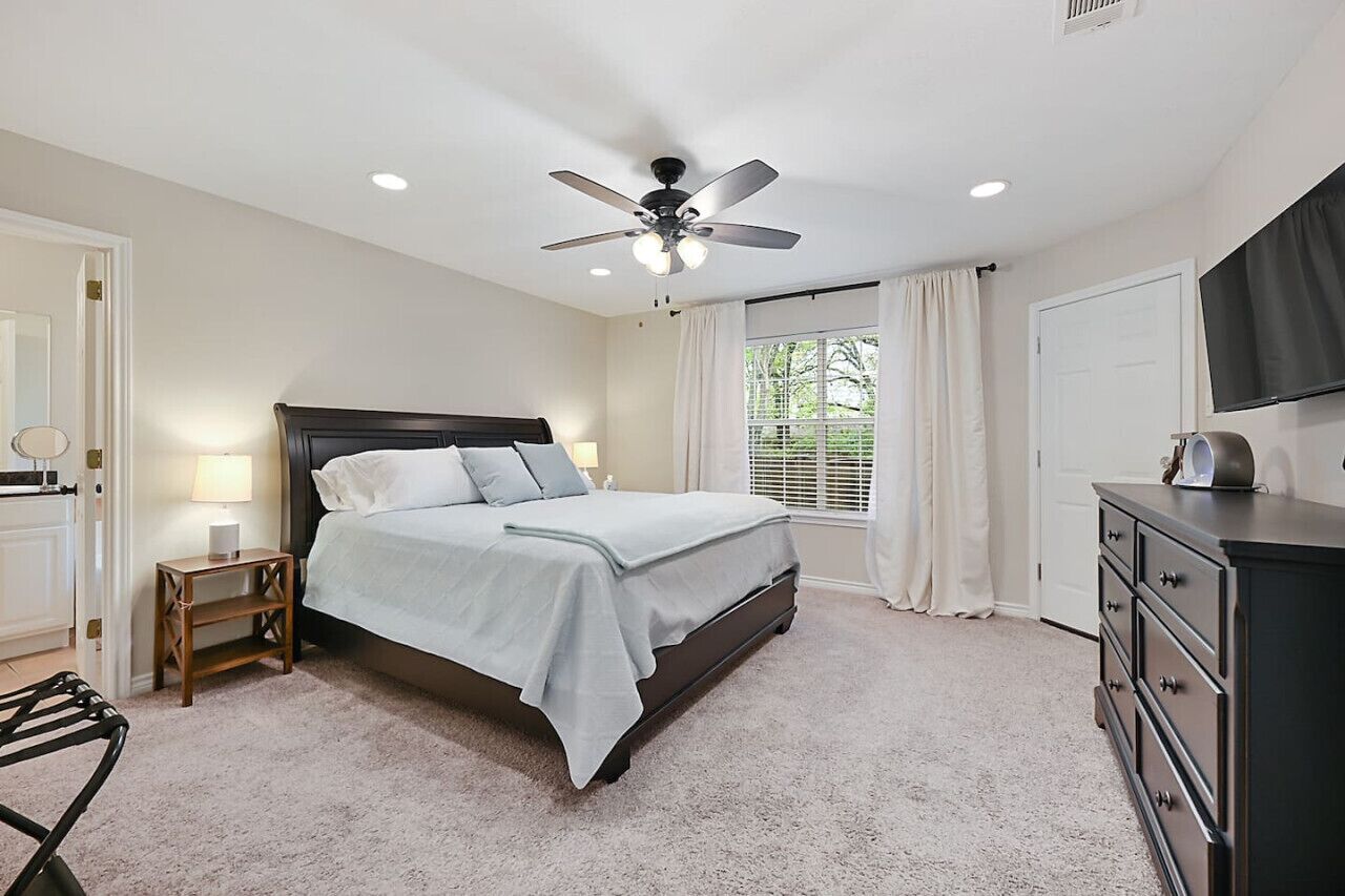Une chambre à coucher comprenant un lit, une table de chevet, une commode, un ventilateur de plafond et une fenêtre avec des rideaux.