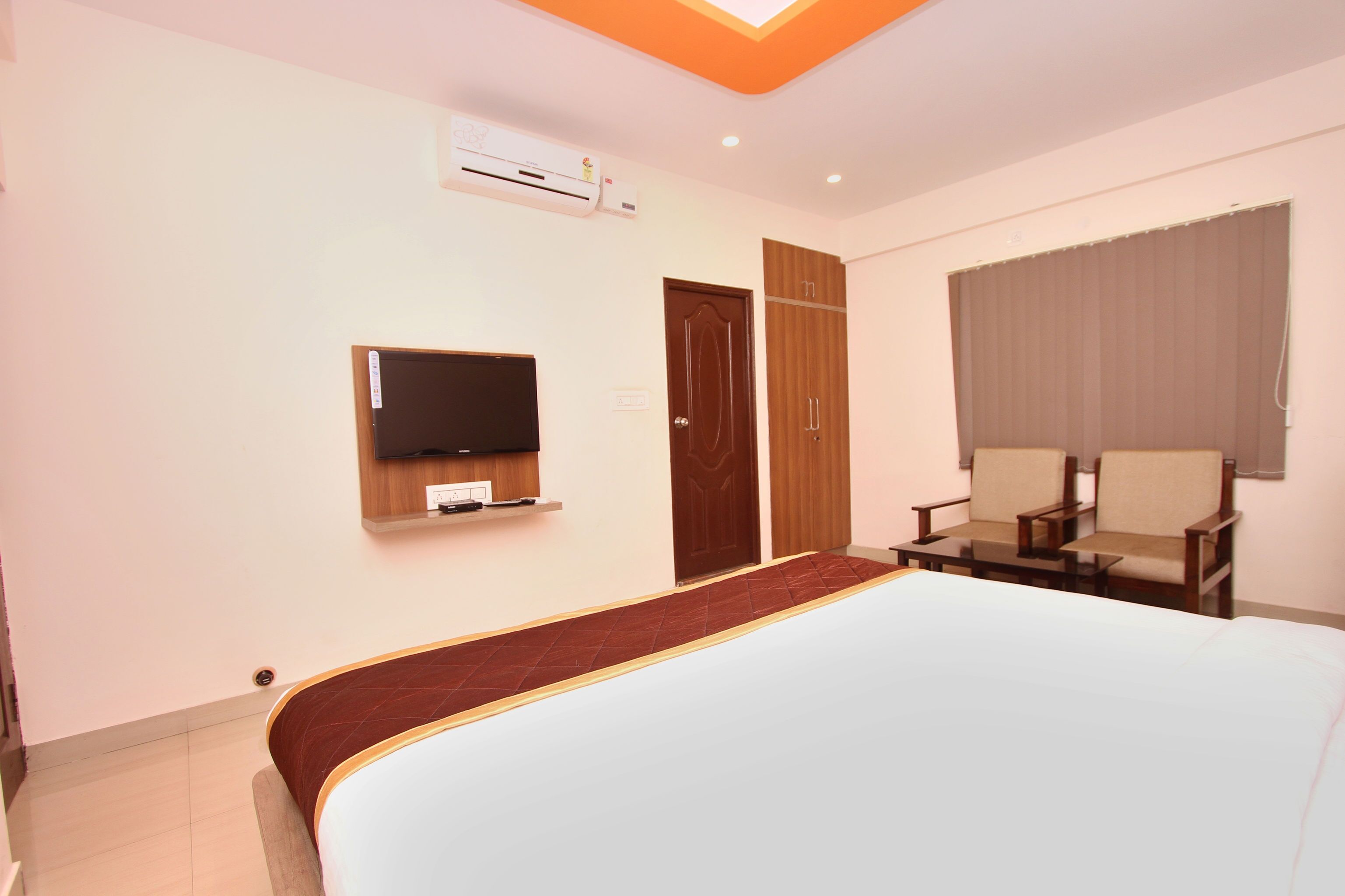 A hotel room with a large bed, a TV mounted on the wall, a wardrobe, a seating area with two chairs, and a window with curtains.