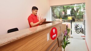 Taulell de recepció amb un home amb samarreta vermella, un mostrador de fusta, un senyal vermell d'OYO Rooms i vista del carrer.