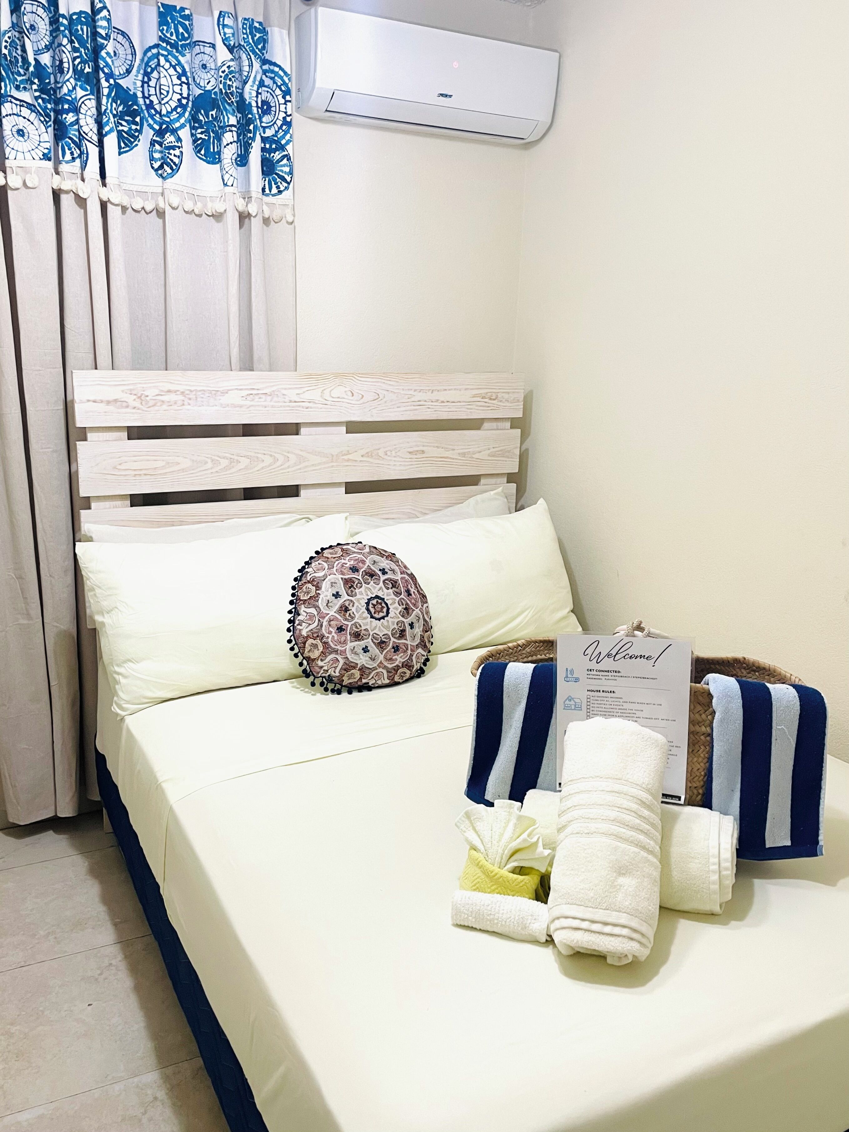 A neatly made bed with a white and blue striped towel, a decorative pillow, and a welcome sign.