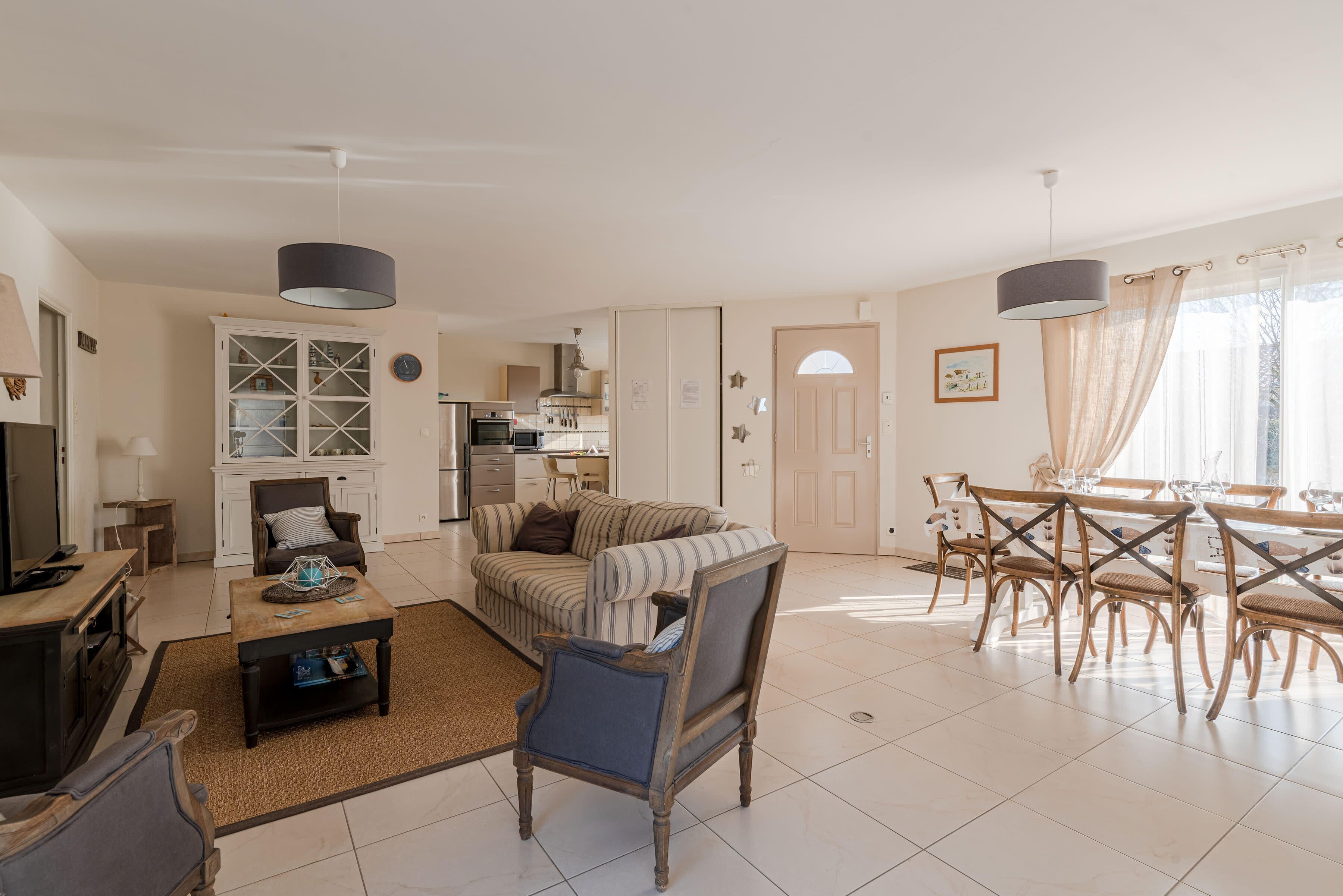 A spacious living area with a sofa, armchairs, a dining table with chairs, a kitchen in the background, and a large window with curtains.