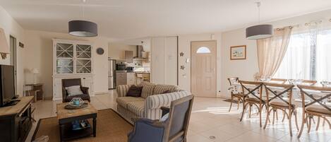 A spacious living area with a sofa, armchairs, a dining table with chairs, a kitchen in the background, and a large window with curtains.