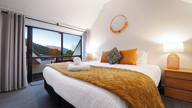 A hotel room with a large bed, a view through a sliding door, a wooden nightstand, and a wall-mounted lamp. - Modern and Convenient apartment in Fernhill (Queenstown)