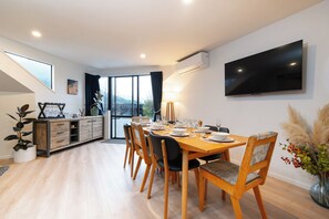 Uma área de refeições moderna com mesa de madeira posta para quatro pessoas, rodeada por cadeiras, num espaço com uma grande janela e televisão de tela plana na parede.