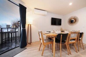 A dining area with a table set for four, a flat-screen TV mounted on the wall, and a view of mountains through a large window. - Sarahs Scenic Stay | Mountain Lake Views (Queenstown)