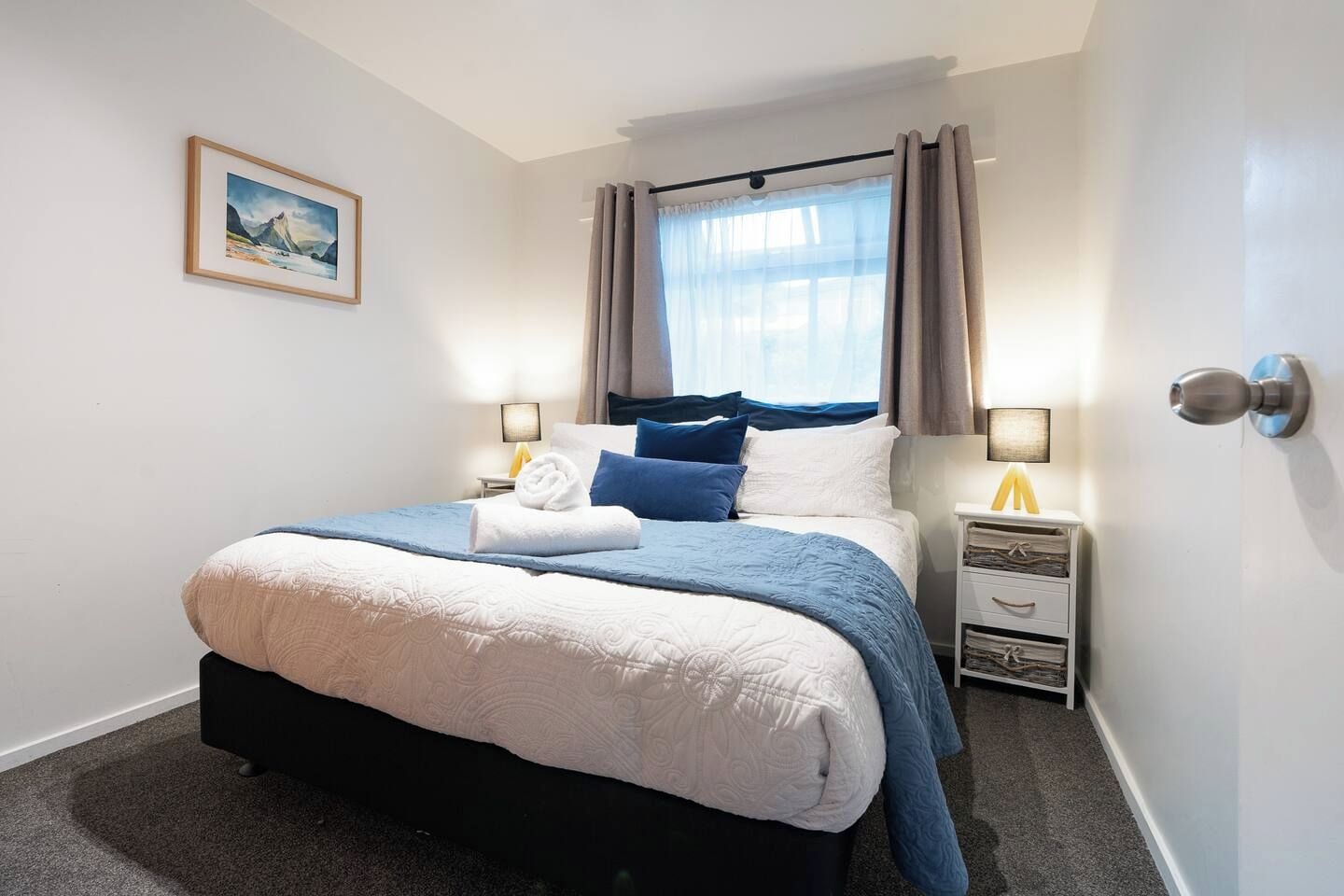 Une chambre d’hôtel avec un lit, une table de chevet, une lampe, un tableau et une fenêtre avec des rideaux.