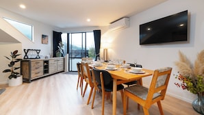 A dining area with a wooden table set for six, surrounded by chairs with cushioned seats. A large flat-screen TV is mounted on the wall above the table. - Modern and Convenient apartment in Fernhill (Queenstown)